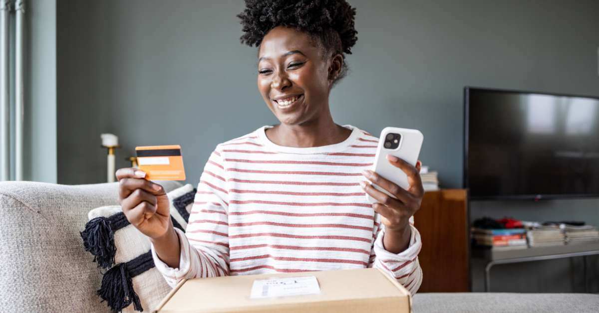 Woman smiles looking at a credit card in her right hand and holding her phone in her left hand while a package she ordered using an app is on her lap.