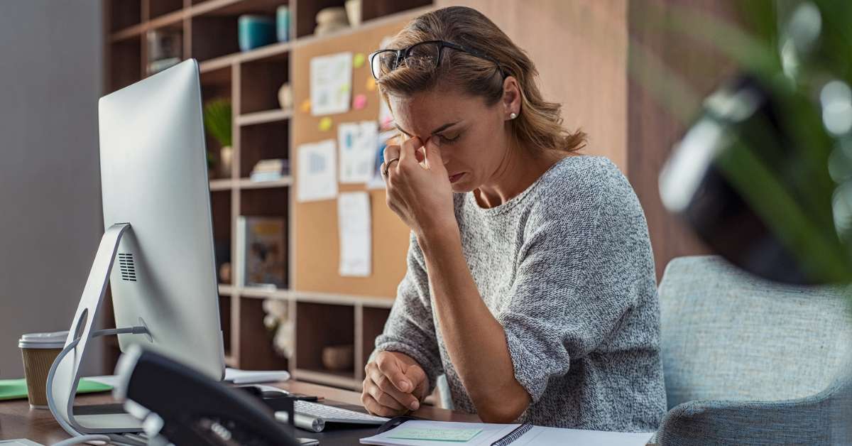 Woman sits at a work desk holding her head out of frustration due to exhaustion from daylight savings time sleep loss.